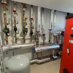 Kompleksowa instalacja grzewcza w kotłowni: rzędy rur w izolacji, manometry, zawory, pompy obiegowe Grundfos, zbiornik wyrównawczy szary, duży czerwony zasobnik ciepłej wody i kratka ściekowa...