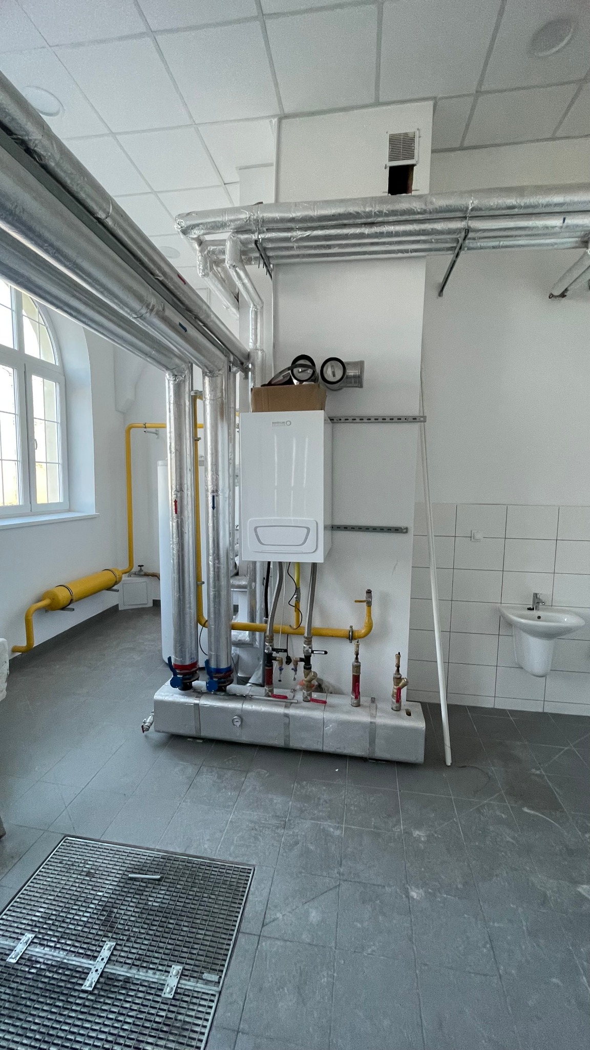 Biały wiszący piec gazowy z podłączonymi rurami i zaworami, zamontowany na ścianie z szarą kratką wentylacyjną w pomieszczeniu z instalacjami grzewczymi i sanitarnymi.