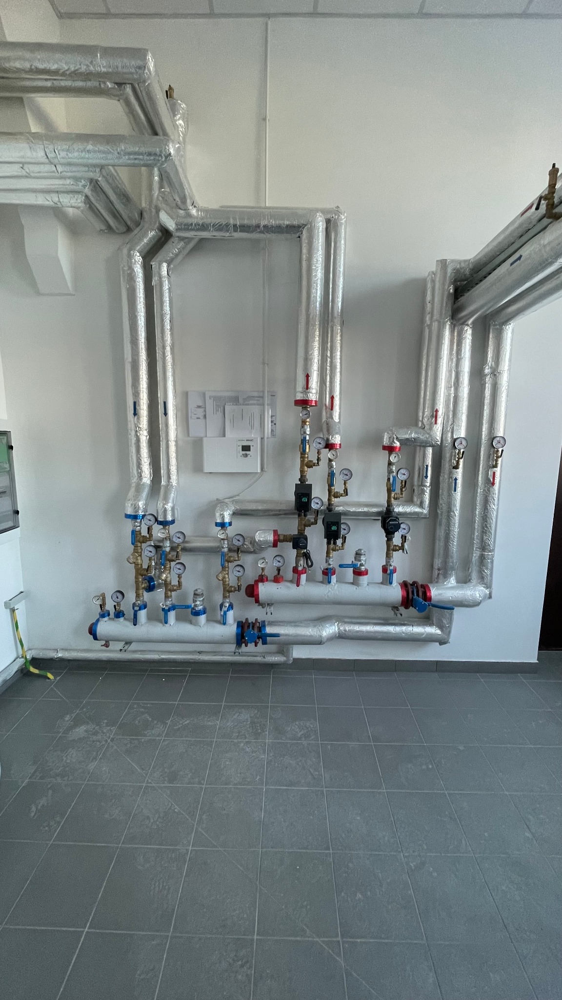 Rozbudowana instalacja rur w izolacji aluminiowej z licznymi zaworami, manometrami i czujnikami, zamontowana na białej ścianie w pomieszczeniu z szarą podłogą z płytek.