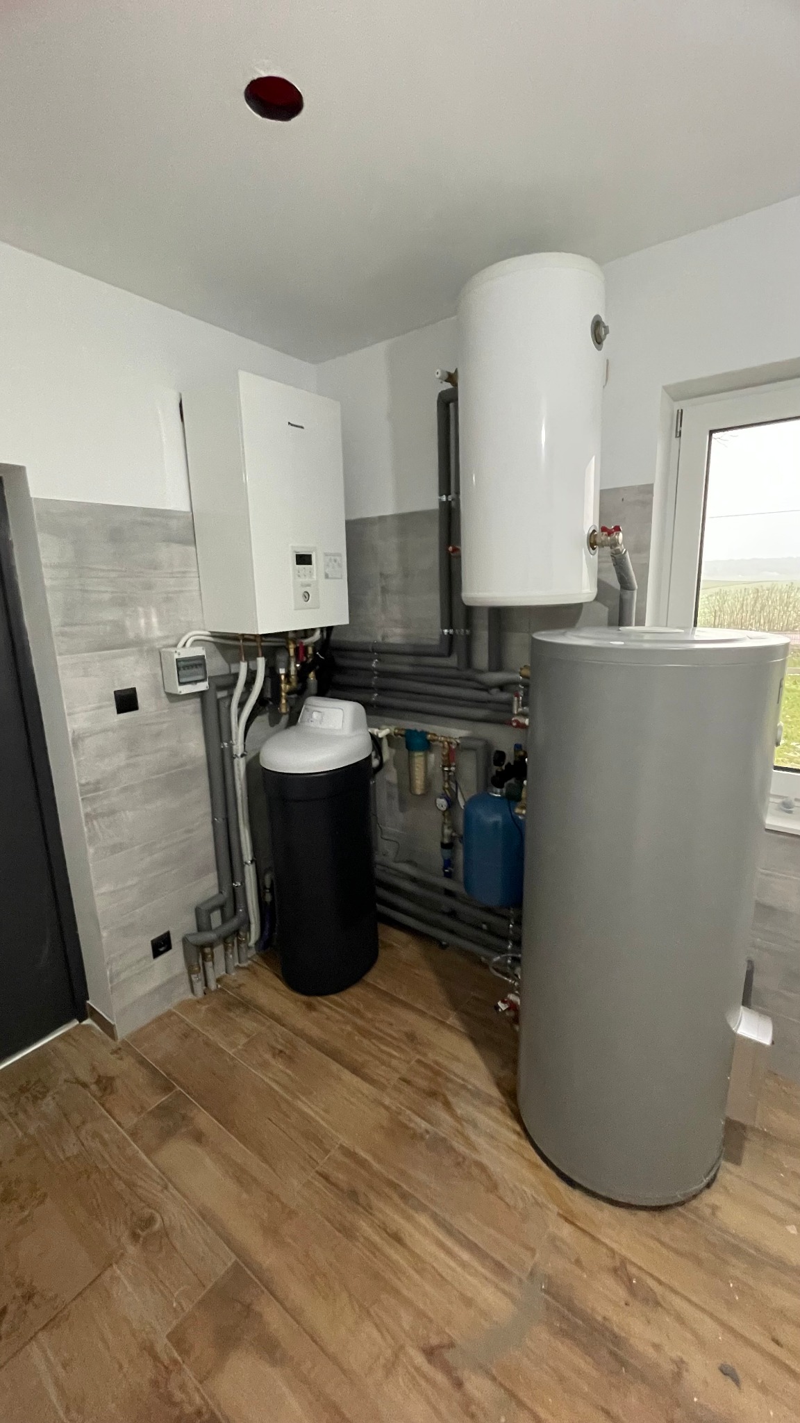 Kotłownia w jasnych barwach z piecem gazowym, zbiornikiem buforowym, zmiękczaczem wody i rozbudowaną instalacją hydrauliczną z licznymi rurami i zaworami.