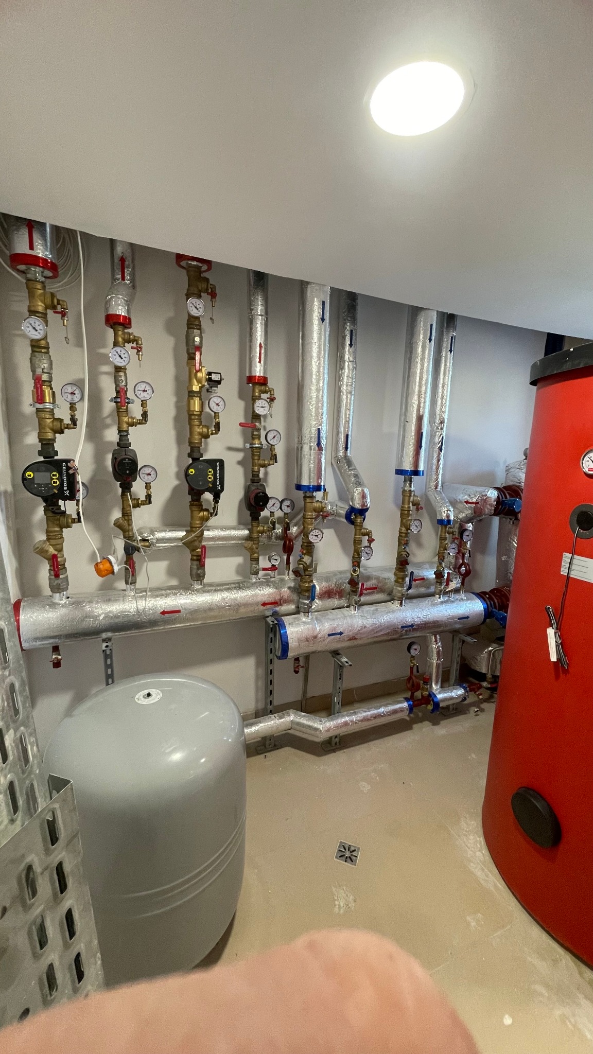 Kompleksowa instalacja grzewcza w kotłowni: rzędy rur w izolacji, manometry, zawory, pompy obiegowe Grundfos, zbiornik wyrównawczy szary, duży czerwony zasobnik ciepłej wody i kratka ściekowa...