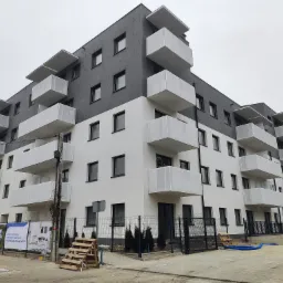 Nowoczesny budynek mieszkalny z szarym i białym wykończeniem, balkonami z ażurowymi balustradami, widok z poziomu ulicy na pochmurny dzień.