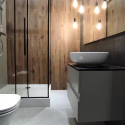 Nowoczesna łazienka z kabiną prysznicową z czarnymi profilami, umywalką nablatową na szafce z szufladami i ścianą wyłożoną panelami imitującymi drewno.