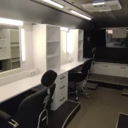 Wnętrze mobilnej garderoby lub wizażowni z białymi meblami na wymiar, lustrami z oświetleniem, czarnymi krzesłami i widokiem na zewnątrz przez okno.