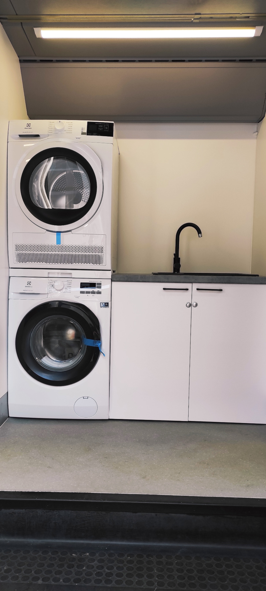 Ustawiona pionowo pralka i suszarka Electrolux w jasnym pomieszczeniu z szafkami pod blatem i czarną baterią.