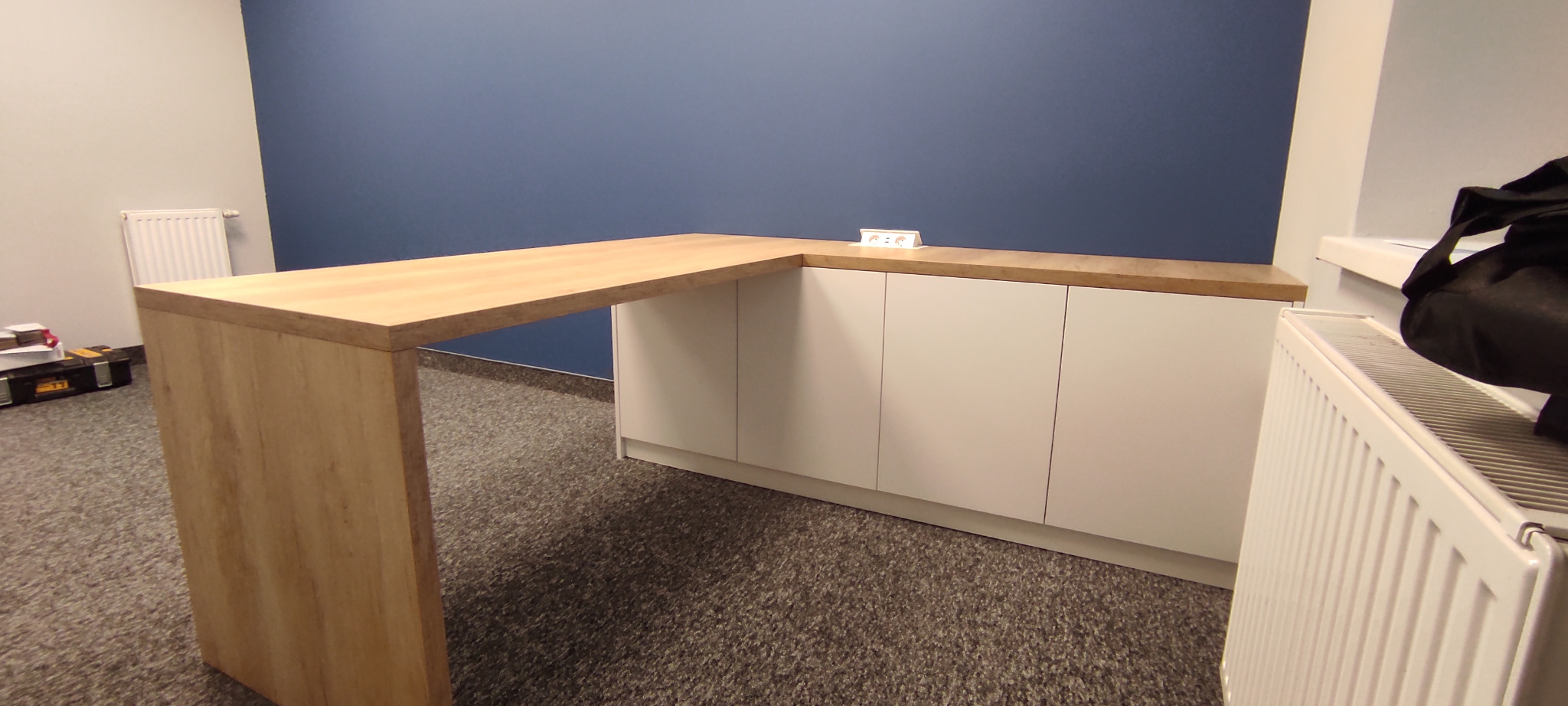 Blat drewniany w kształcie litery L, połączony z białymi szafkami, na tle niebieskiej ściany. Na blacie widoczny biały przedłużacz.