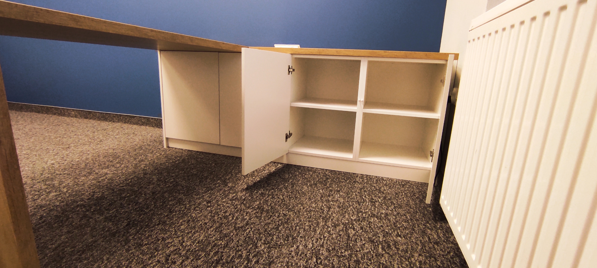 Biała szafka z otwartymi drzwiczkami, z drewnianym blatem, na tle niebieskiej ściany i dywanu w odcieniach szarości.