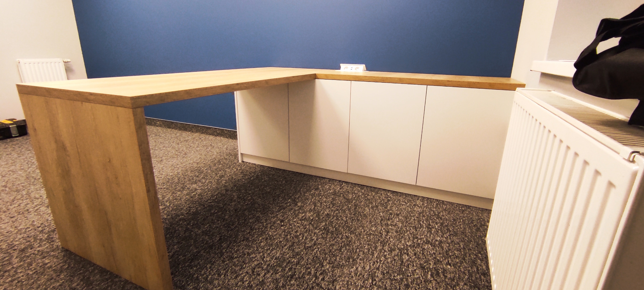 Kątowy blat drewniany o jasnym odcieniu drewna, połączony z białymi szafkami z frontami bez uchwytów, na tle niebieskiej ściany. Widoczny fragment grzejnika i skrzynka narzędziowa.