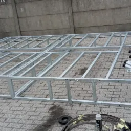Szkielet metalowej konstrukcji z profili, prawdopodobnie przygotowywanej do montażu, leżący na brukowanej powierzchni, z widocznymi elementami montażowymi i oponami w tle.