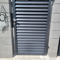 Nowoczesna, grafitowa brama wjazdowa z poziomymi lamelami, zamontowana między betonowymi słupkami z widocznym okablowaniem po lewej stronie.