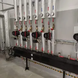 Rozbudowana instalacja grzewcza z pompami obiegowymi Grundfos, rurami w izolacji aluminiowej i manometrami, zamontowana w jasnym pomieszczeniu technicznym.