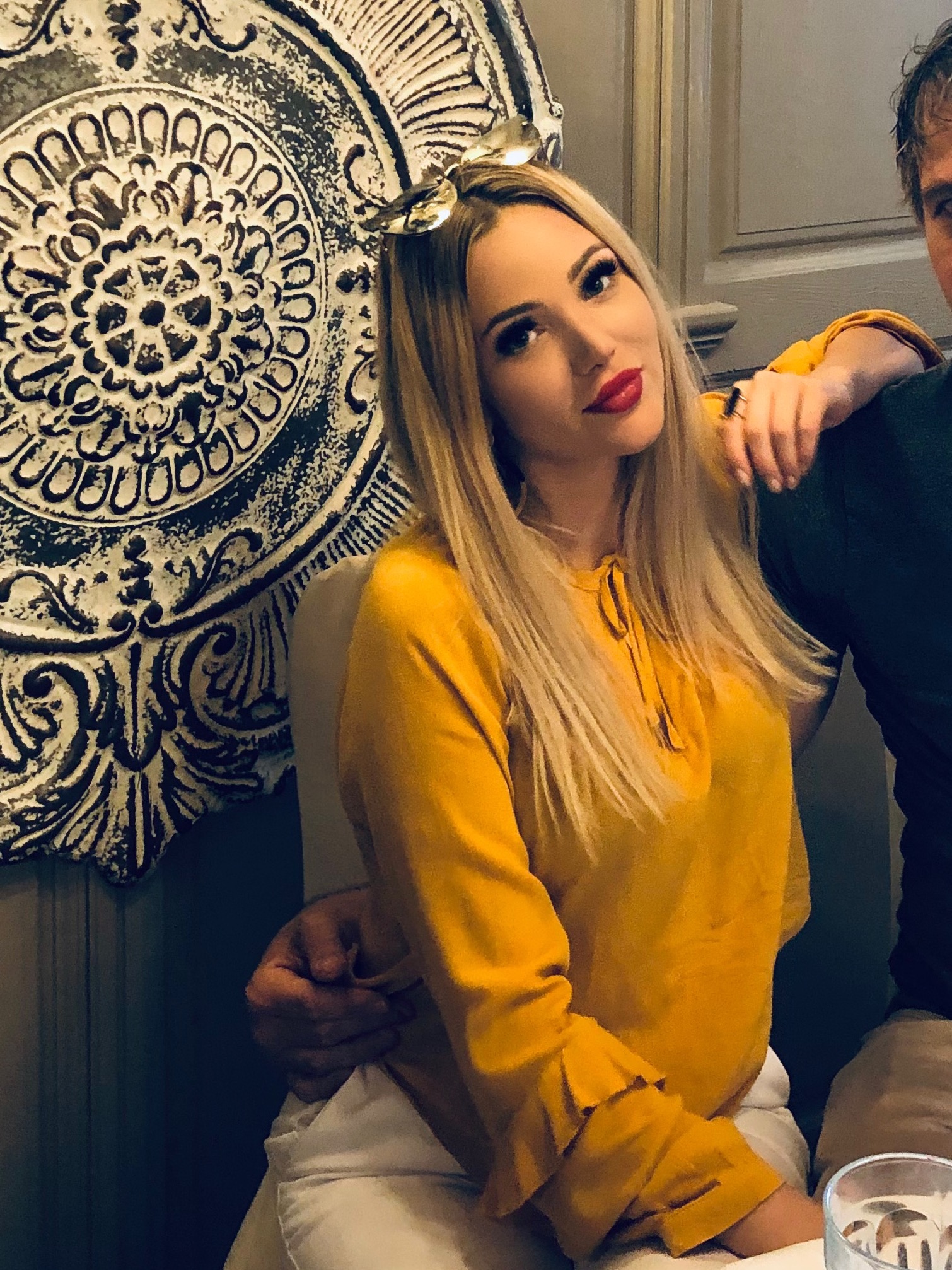 Portret kobiety w żółtej bluzce z kokardą, siedzącej obok mężczyzny, na tle ozdobnej ściany.