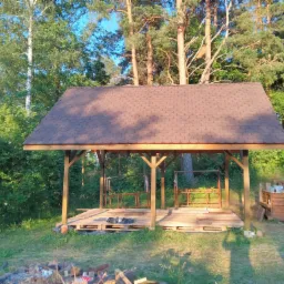 Drewniana altana z brązowym dachem z gontu bitumicznego w trakcie budowy, posadowiona na paletach, otoczona zielenią drzew i trawy.