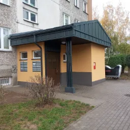 Odnowione wejście do budynku wielorodzinnego z nowym zadaszeniem w kolorze grafitowym, żółtą elewacją i skrzynkami pocztowymi, widoczne graffiti na sąsiedniej ścianie i motocykl pod pokrowcem.