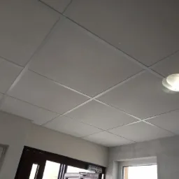 Kasetonowy sufit podwieszany z oświetleniem, widok z dołu, fragment okna i drzwi w tle.