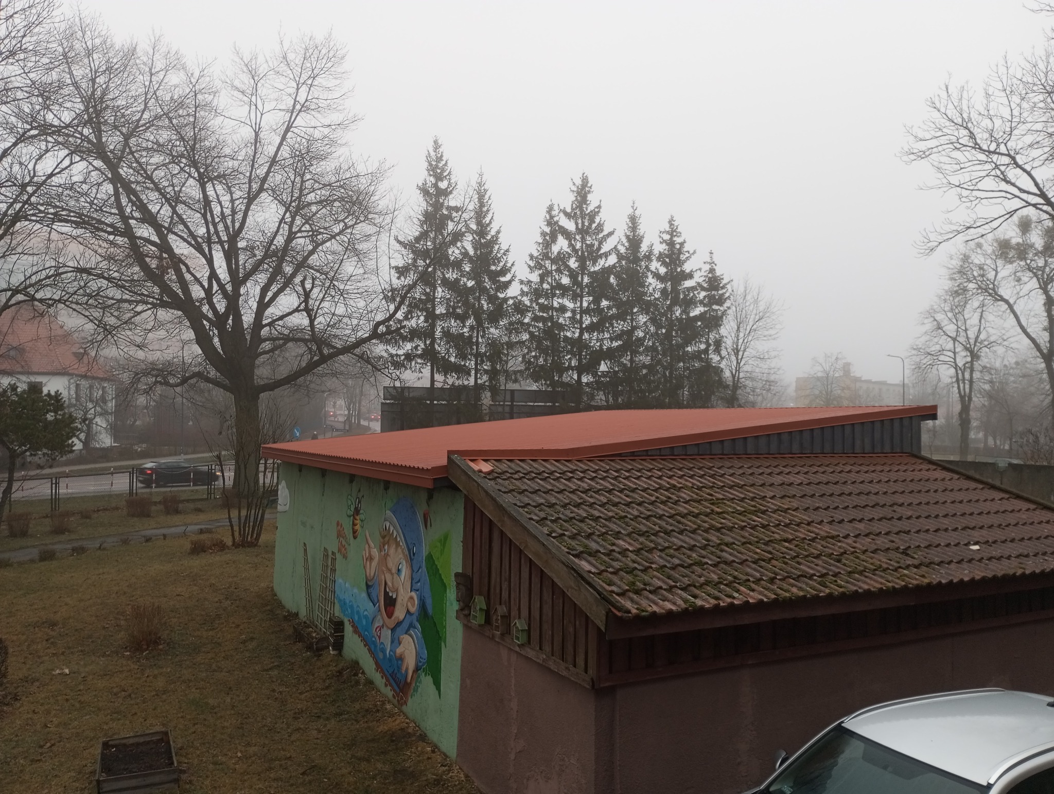 Widok na dwa dachy: stary, pokryty brązową dachówką porośniętą mchem oraz nowy, z blachy w kolorze ceglastym, w tle drzewa i mglisty krajobraz.