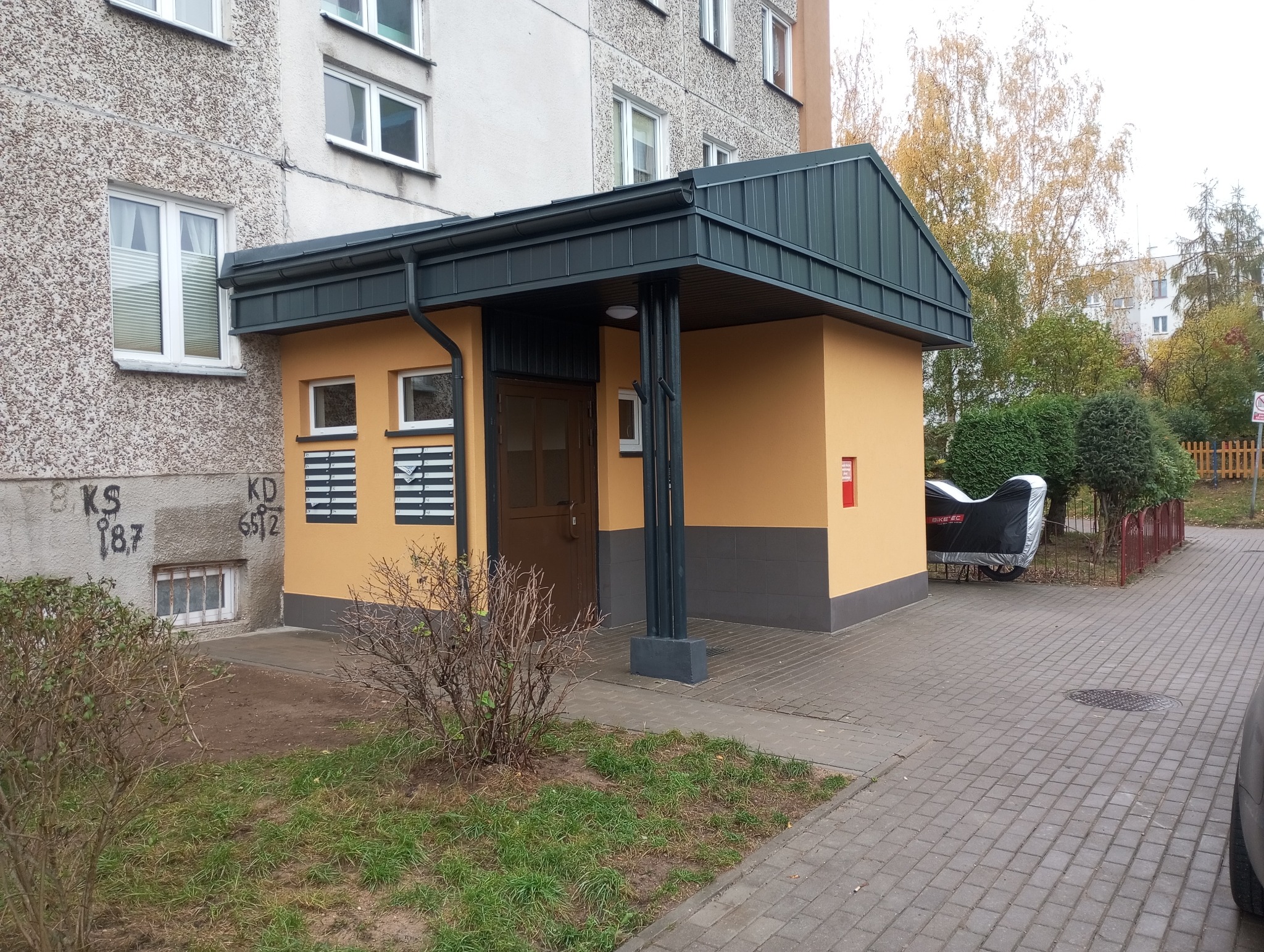 Odnowione wejście do budynku wielorodzinnego z nowym zadaszeniem w kolorze grafitowym, żółtą elewacją i skrzynkami pocztowymi, widoczne graffiti na sąsiedniej ścianie i motocykl pod pokrowcem.