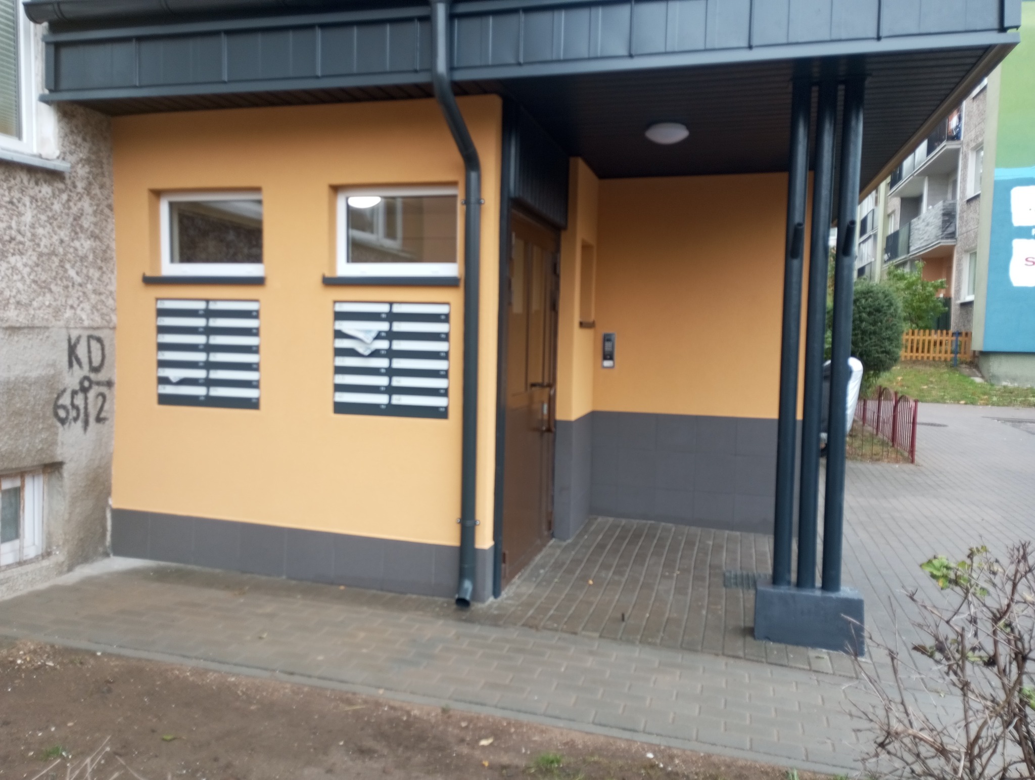 Wejście do budynku z żółtą elewacją, skrzynkami pocztowymi, szarą podmurówką i zadaszeniem wspartym na czarnych kolumnach. Widoczny fragment chodnika i zieleni w tle.