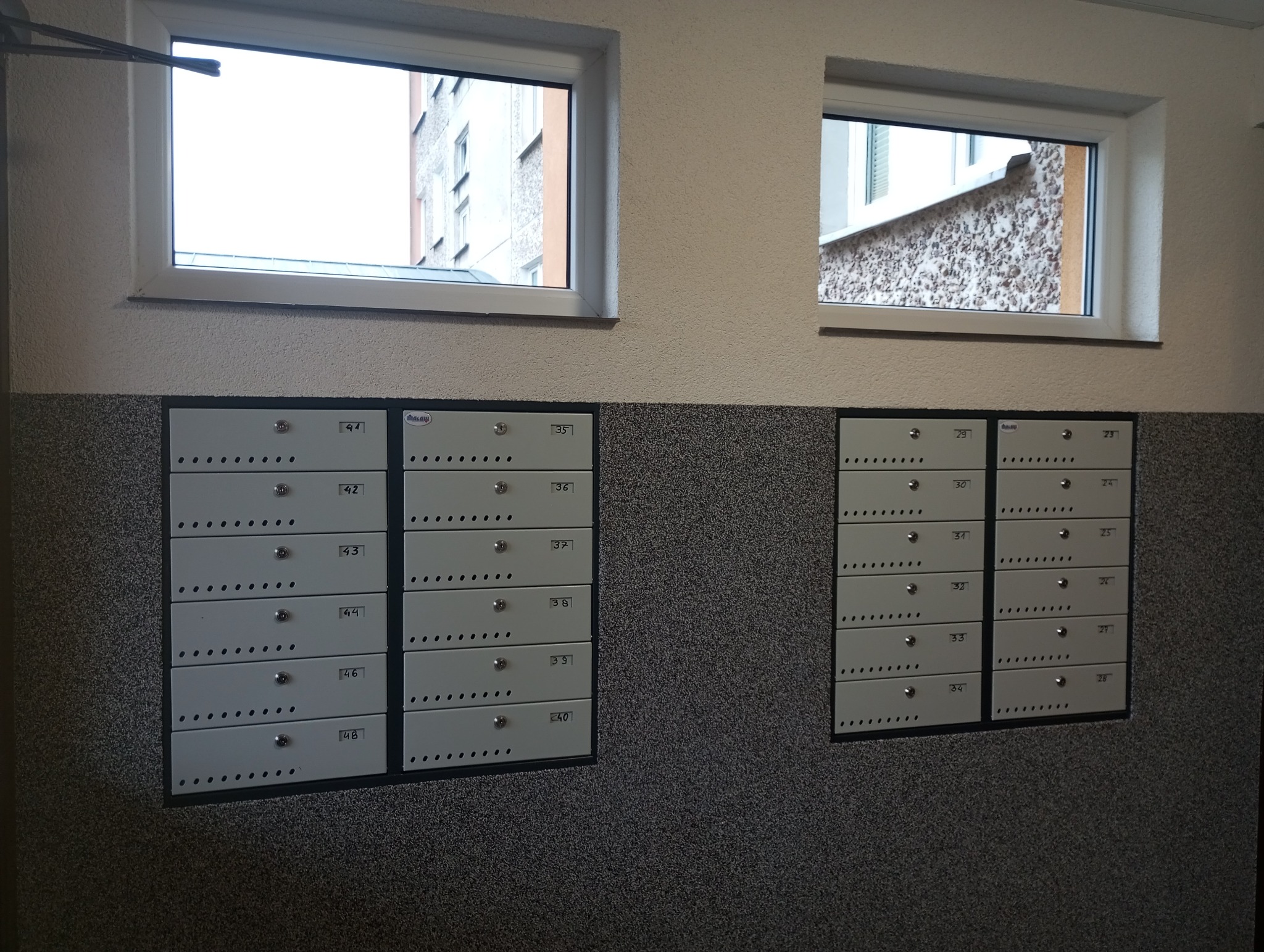 Zestaw skrzynek pocztowych zamontowanych na ścianie wewnątrz budynku, widoczne dwa okna z widokiem na zewnątrz.