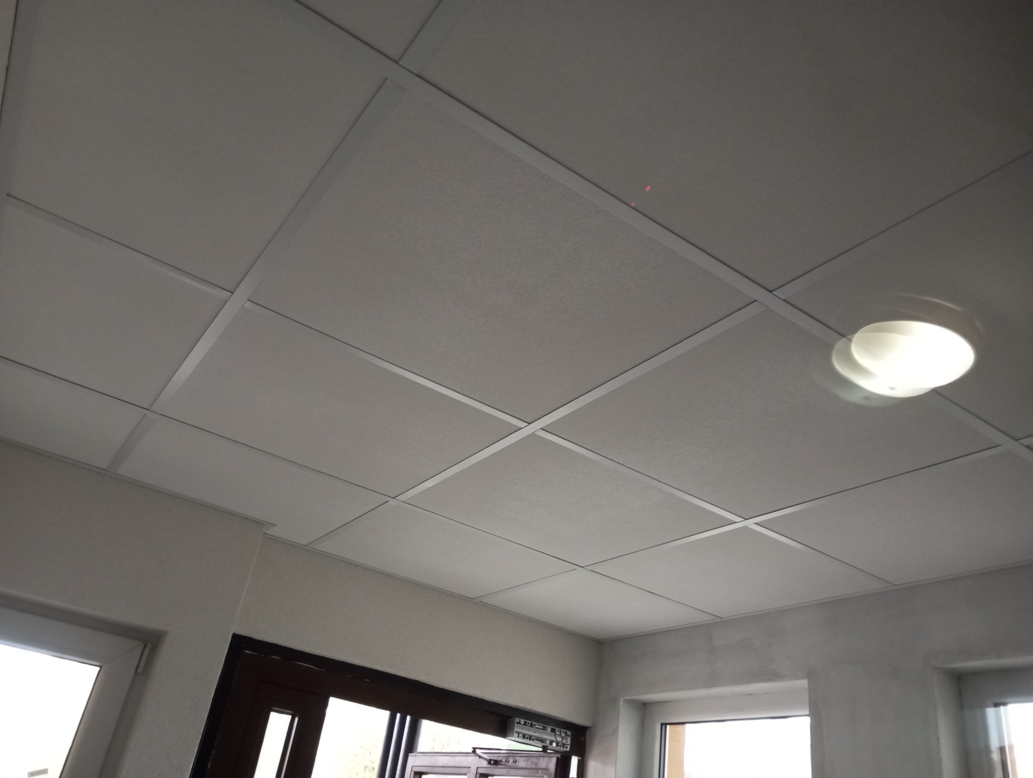 Kasetonowy sufit podwieszany z oświetleniem, widok z dołu, fragment okna i drzwi w tle.