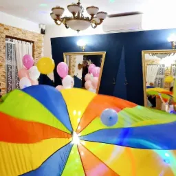 Grupa dzieci i dorosłych bawi się kolorową chustą animacyjną w pomieszczeniu udekorowanym balonami i oświetleniem dyskotekowym.