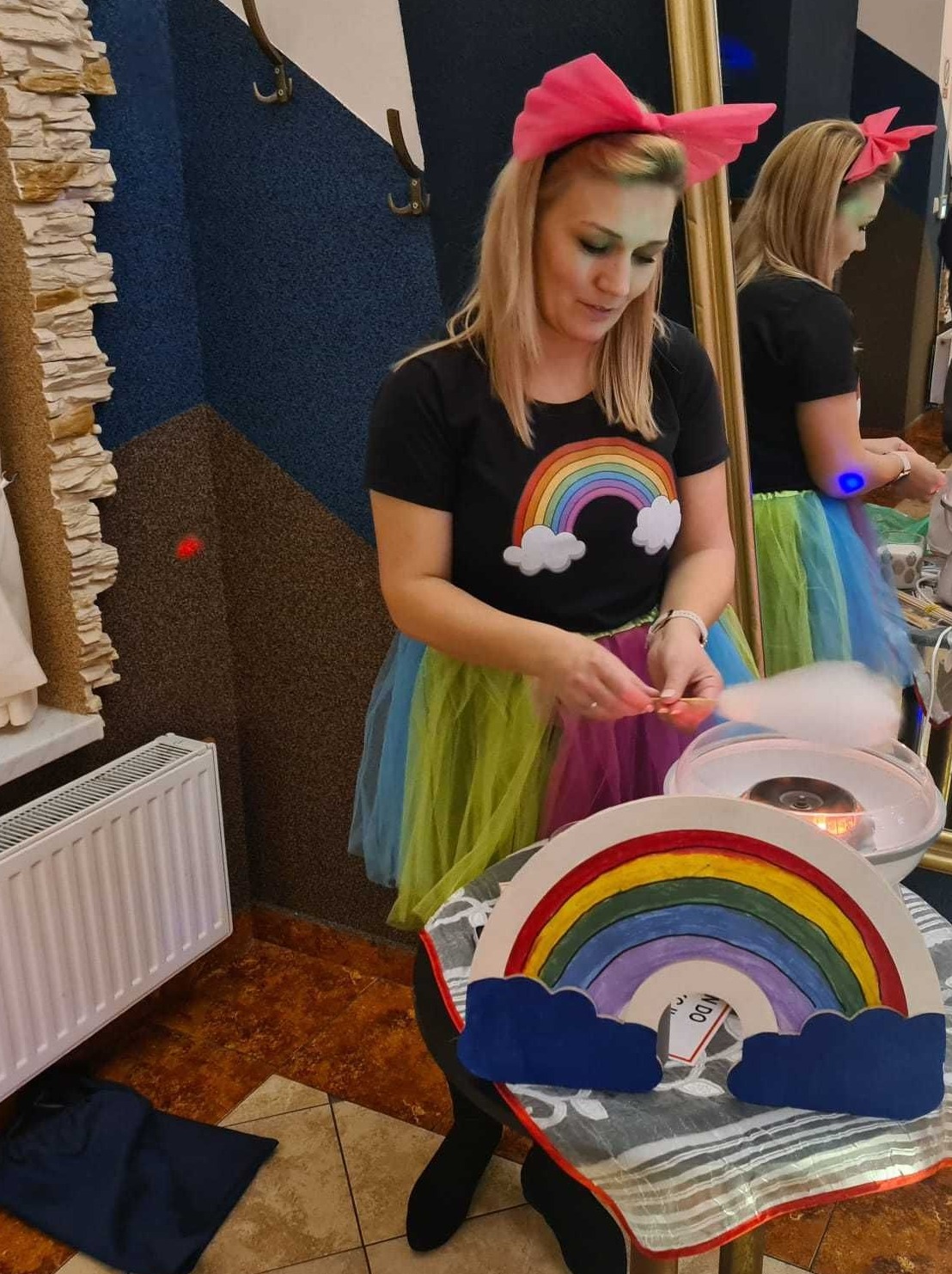 Kobieta w Częstochowie ubrana w tęczową koszulkę i tiulową spódnicę, z różową kokardą na głowie, przygotowuje watę cukrową. Na stole tęczowa dekoracja z chmurkami.