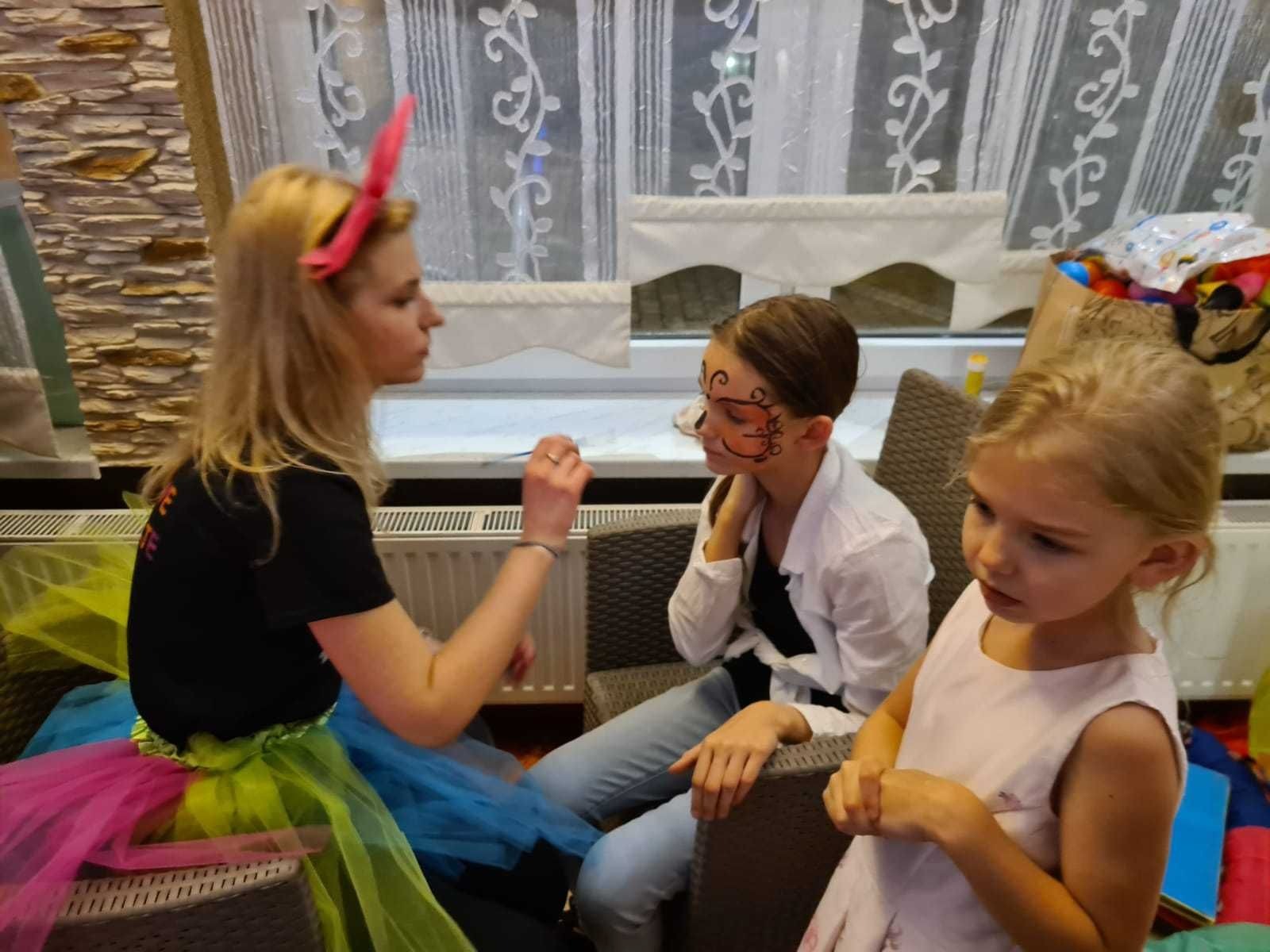 Malowanie twarzy dziewczynce przez animatorkę w kolorowej spódnicy tutu. Druga dziewczynka obserwuje.