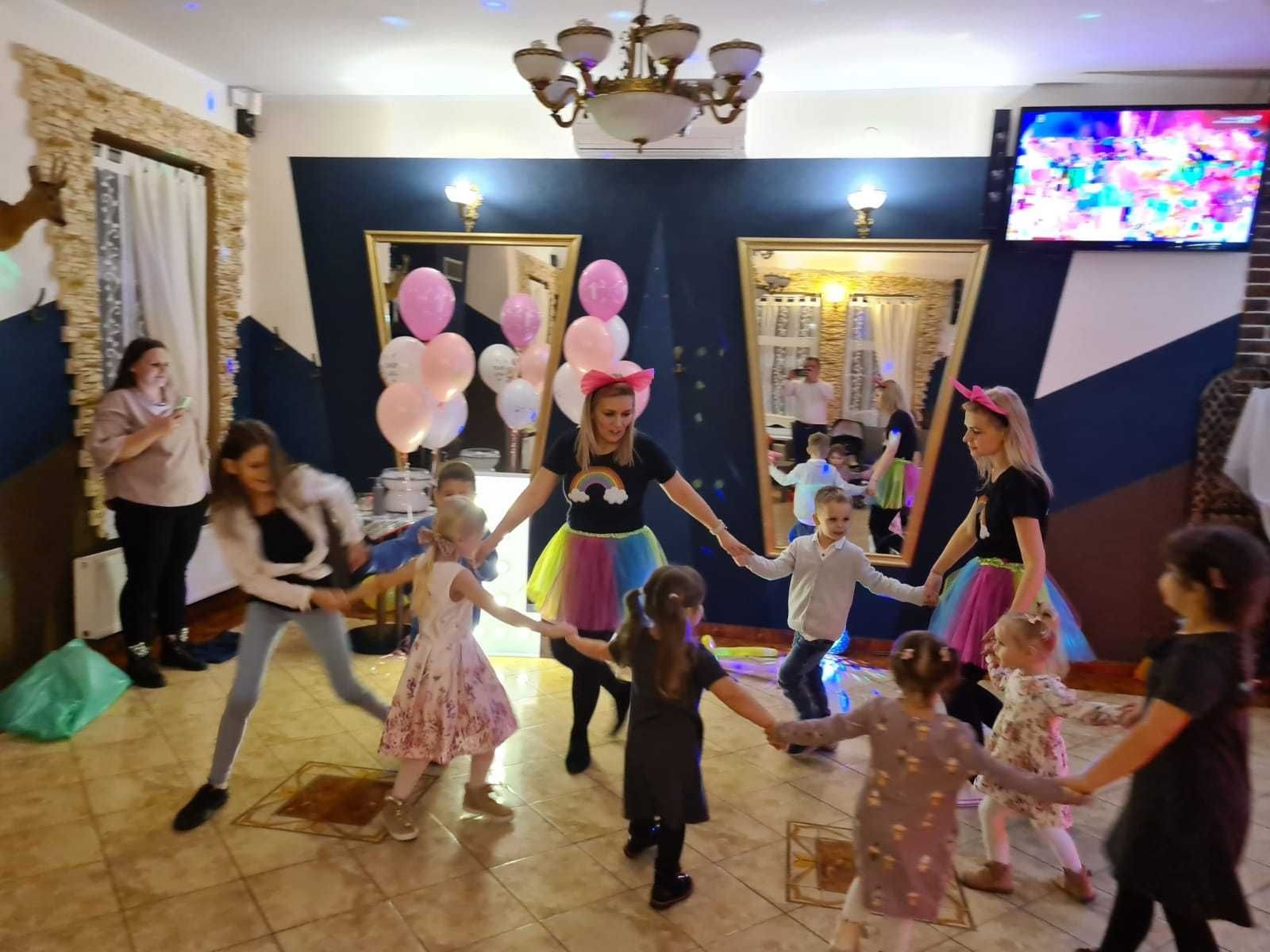 Grupa dzieci trzymających się za ręce, tworzących koło, prowadzona przez kobietę w tęczowej spódnicy i opasce z uszami, w tle balony i lustra w złoconych ramach.