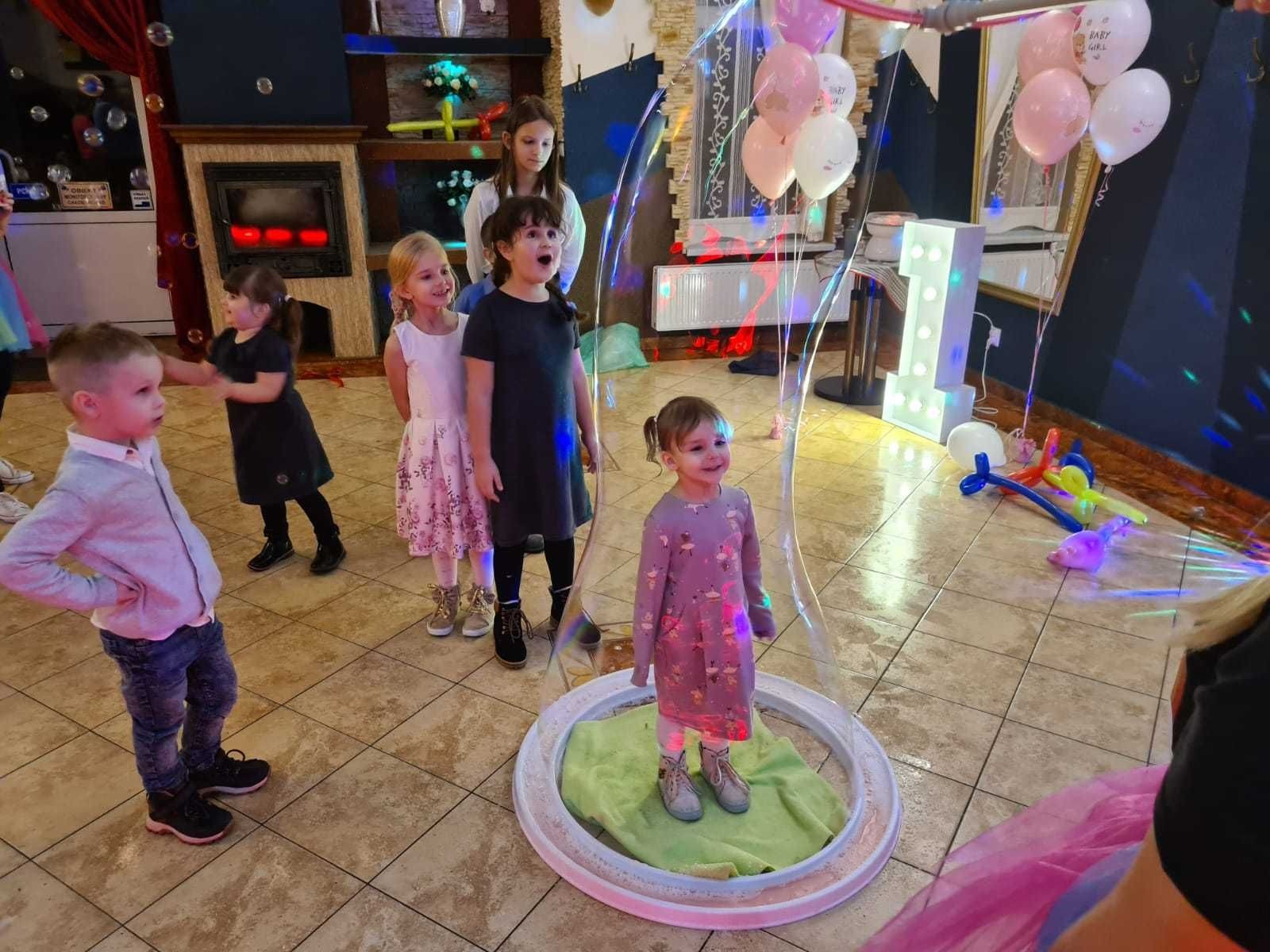 Grupa dzieci w sali zabaw, dziewczynka w fioletowej sukience stoi wewnątrz dużej bańki mydlanej, w tle dekoracje urodzinowe i dyskotekowe światła.