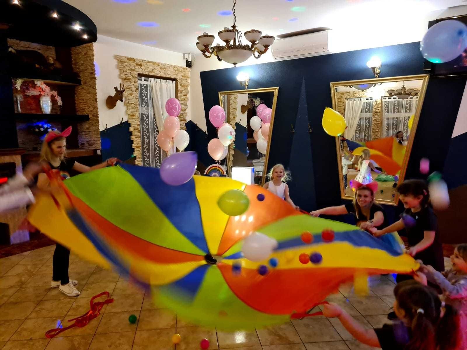 Animatorzy z dziećmi bawią się kolorową chustą animacyjną w sali z dekoracjami balonowymi i oświetleniem dyskotekowym.