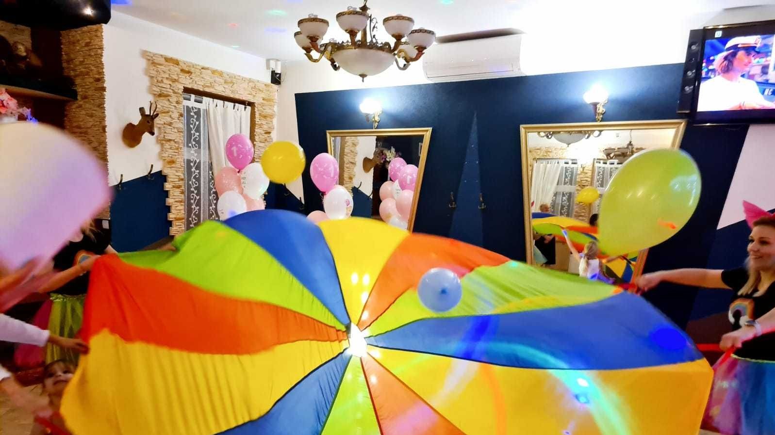 Grupa dzieci i dorosłych bawi się kolorową chustą animacyjną w pomieszczeniu udekorowanym balonami i oświetleniem dyskotekowym.