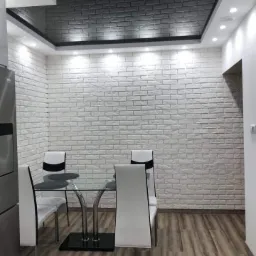 Jadalnia z białą ścianą wyłożoną dekoracyjną cegłą, czarnym sufitem podwieszanym z oświetleniem punktowym, szklanym stołem i białymi krzesłami na podłodze imitującej drewno.