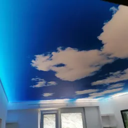 Sufit napinany z motywem nieba i chmur, podświetlony niebieskim światłem LED, widok z dołu na fragment pokoju z oknem i obrazami na ścianie.