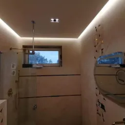 Nowoczesna łazienka z beżowymi płytkami, podświetlanym sufitem, kabiną prysznicową ze złotą armaturą i owalnym lustrem z podświetleniem.