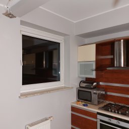 Rafał - Wnętrze kuchni po odświeżeniu, z widocznym oknem z roletą, grzejnikiem, zabudową kuchenną z drewnianymi frontami, kuchenką gazową i mikrofalówką.