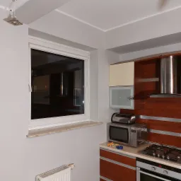 Wnętrze kuchni po odświeżeniu, z widocznym oknem z roletą, grzejnikiem, zabudową kuchenną z drewnianymi frontami, kuchenką gazową i mikrofalówką.