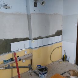 Rafał - Wnętrze w trakcie układania białych, prostokątnych płytek na ścianie, widoczny grunt na ścianie, poziomica, rury, wiadro z gruntem i narzędzia glazurnicze.