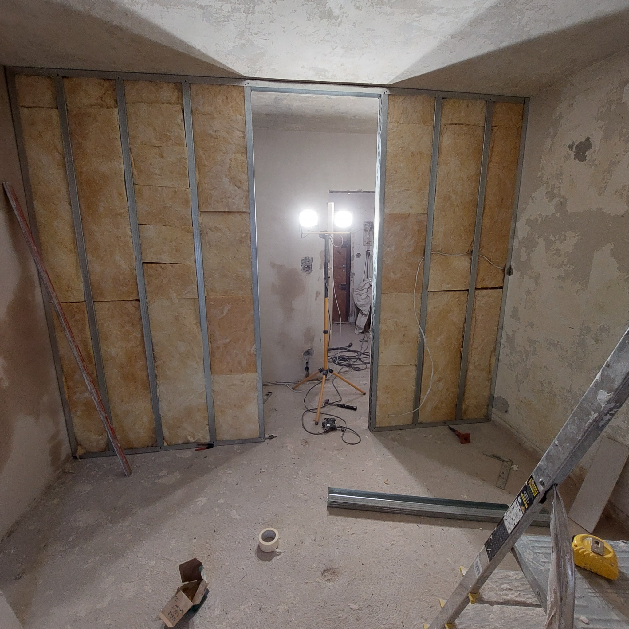 Wnętrze w trakcie prac, widoczne metalowe profile konstrukcyjne z wypełnieniem z wełny mineralnej, oświetlenie budowlane na trójnogu, drabina aluminiowa, surowe ściany i podłoga w trakcie...