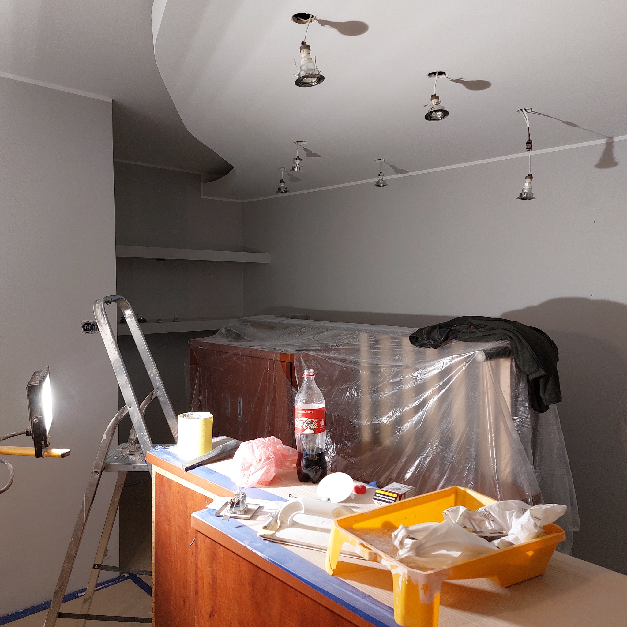 Pokój w trakcie odświeżania, widoczne przygotowania do malowania ścian: drabina, lampa halogenowa, zabezpieczone meble folią, puszka z farbą, taśma malarska i napój Coca-Cola.