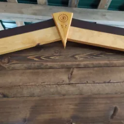 Detal drewnianej konstrukcji z dekoracyjnym elementem w kształcie kwiatu słonecznika, ciemnobrązowe deski i jasne drewno, widoczna część dachu z zieloną blachą.