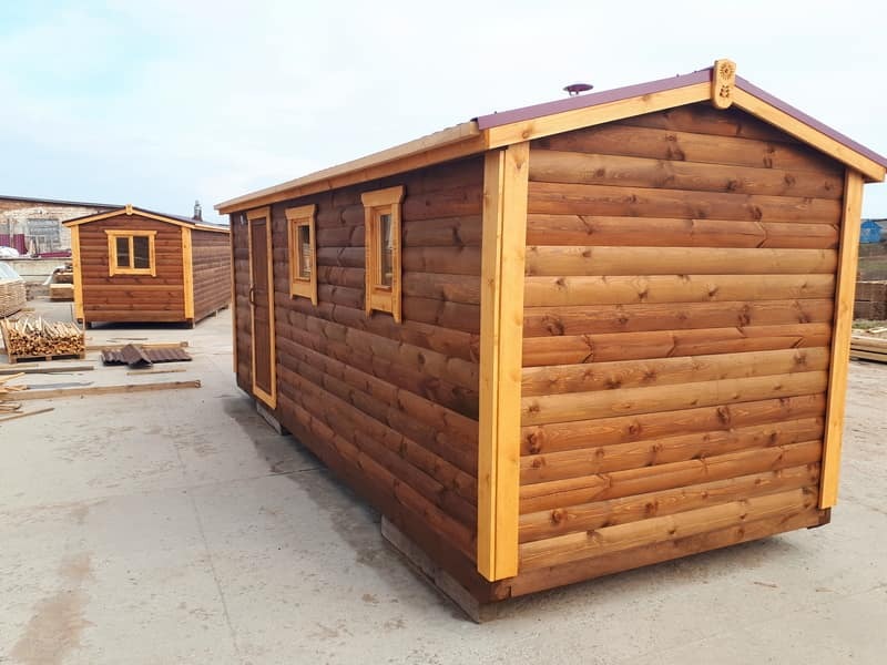 Gotowa, drewniana konstrukcja z bali, prawdopodobnie mobilna, z oknami i drzwiami, z dekoracyjnym wykończeniem dachu, na placu budowy, z drugą podobną konstrukcją w tle i stosem drewna.