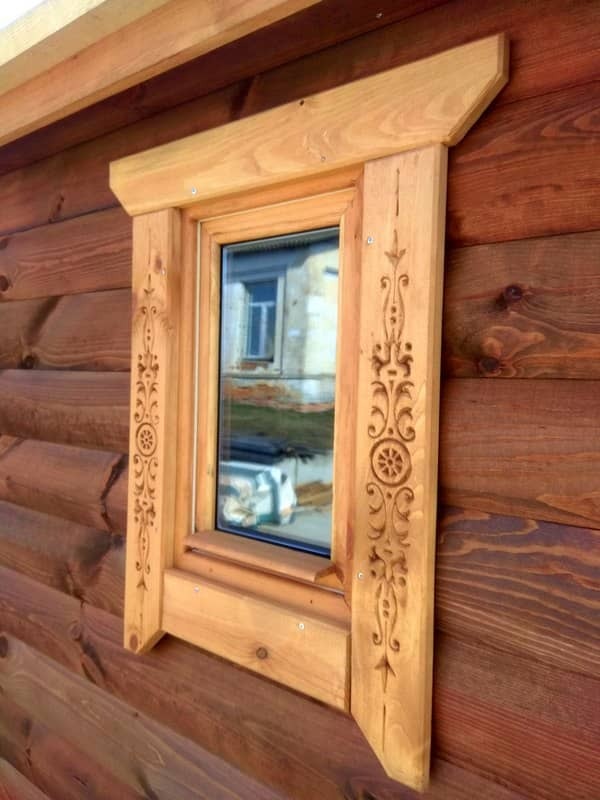Drewniana rama okienna z dekoracyjnymi rzeźbieniami, osadzona w ścianie z bali w odcieniach brązu.