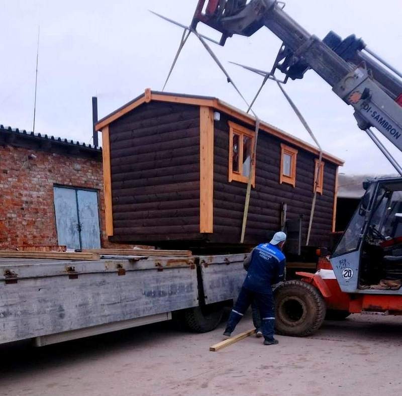Przenośna, ciemnobrązowa sauna z jasnymi obramowaniami okien i dachu, podnoszona przez dźwig z wysięgnikiem, w tle ceglany budynek.