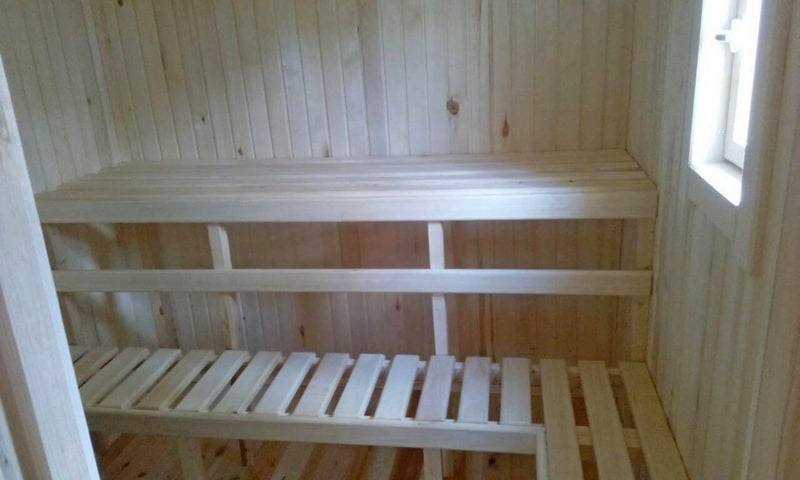 Wnętrze nowej, jasnej, drewnianej kabiny z dwupoziomowymi ławkami, widoczne małe okno.