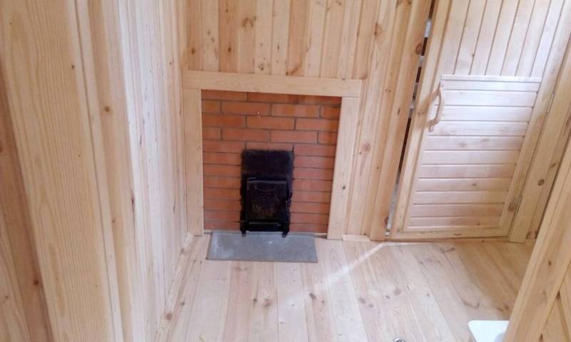 Wnętrze drewnianej kabiny z piecem na drewno, widok na ceglany komin z metalowymi drzwiczkami, jasne drewno na ścianach i podłodze, drzwi z poziomymi panelami po prawej stronie.