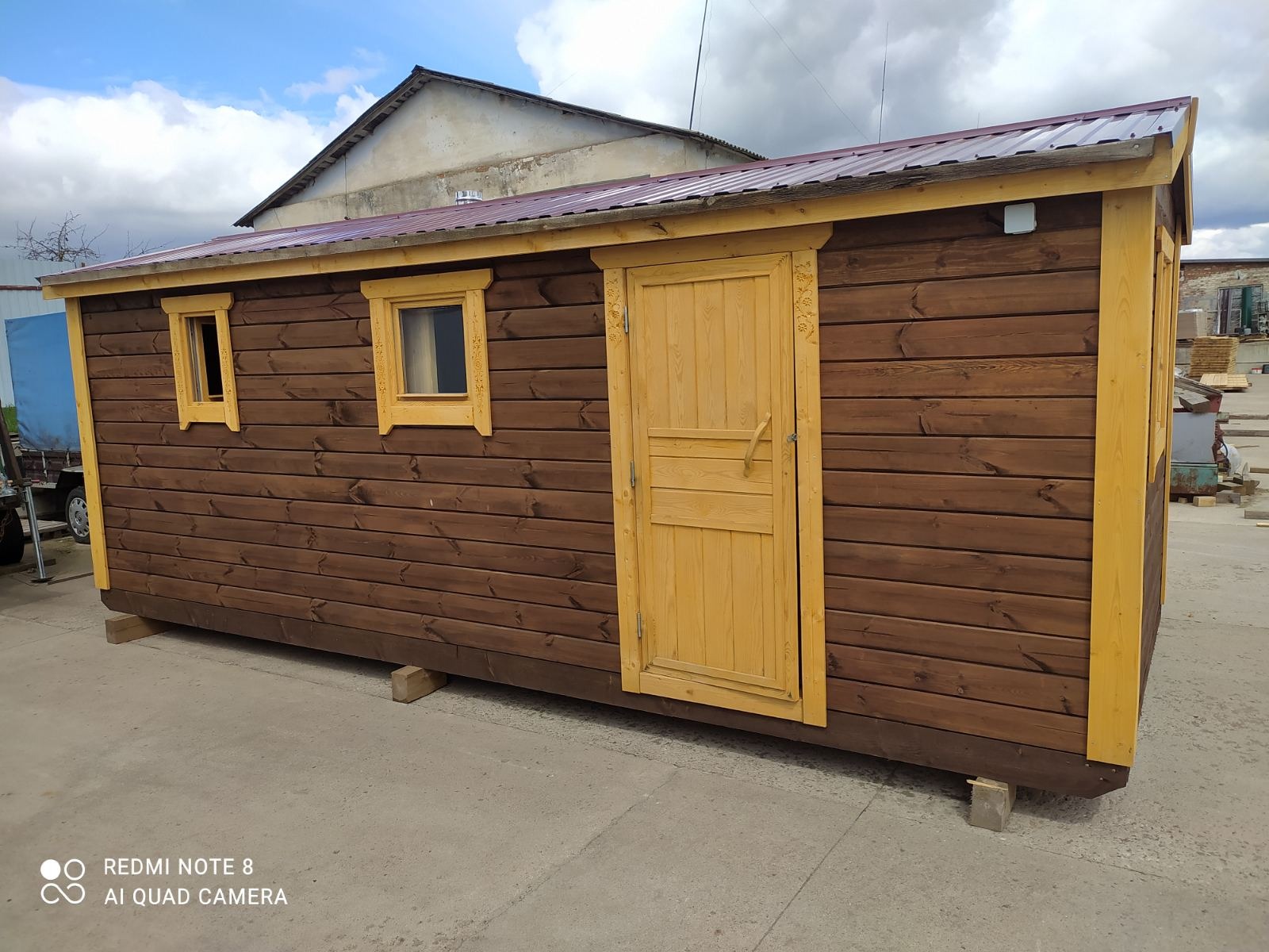 Gotowa, drewniana konstrukcja z dwoma oknami i drzwiami, ustawiona na betonowej powierzchni, z widocznym budynkiem w tle.