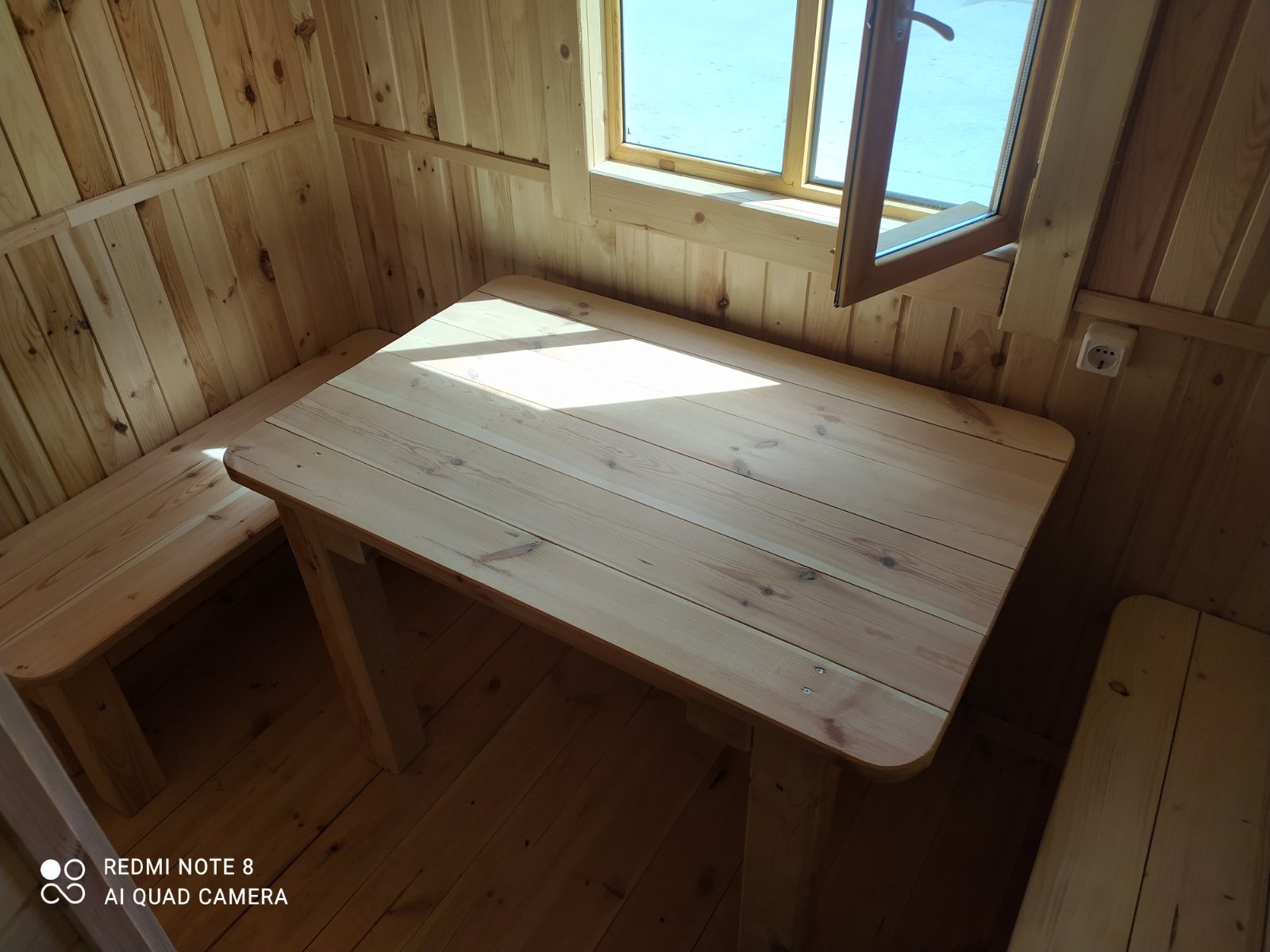 Wnętrze drewnianej kabiny z jasnym stołem i ławkami, widok przez otwarte okno na zewnątrz.