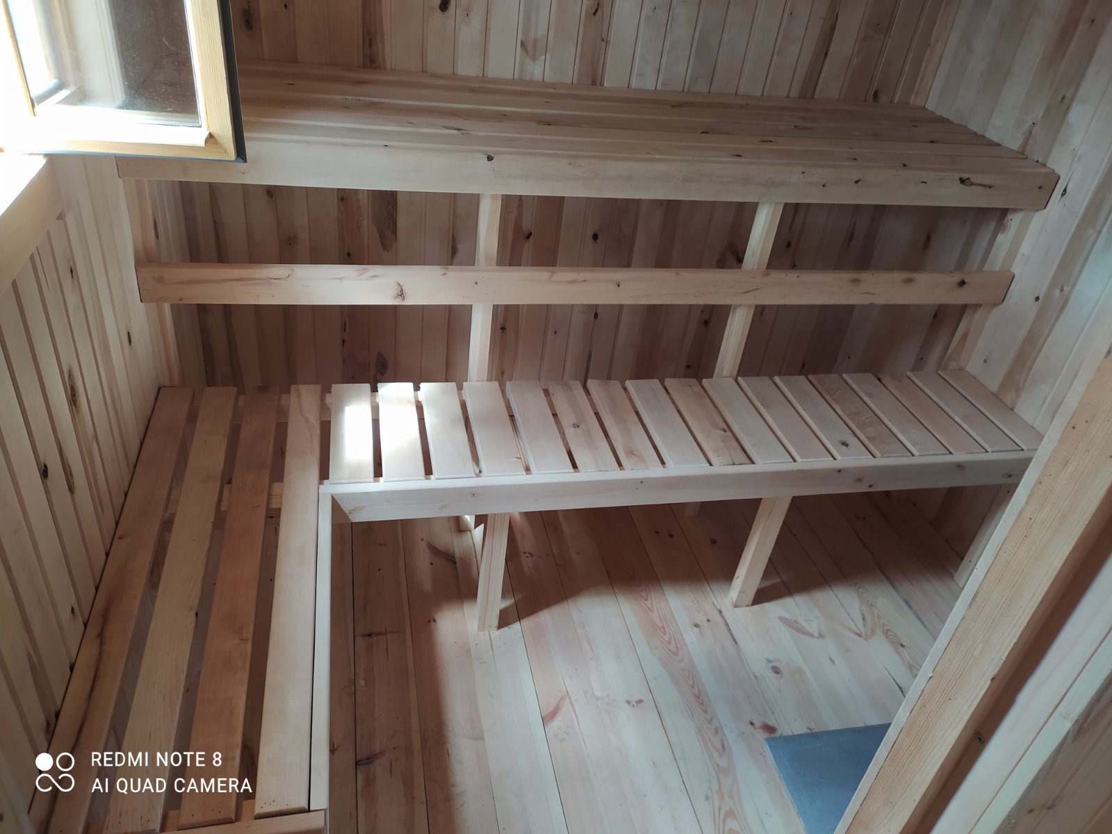 Wnętrze wykończonej drewnianej kabiny z ławkami i półkami, widok na okno w górnym rogu