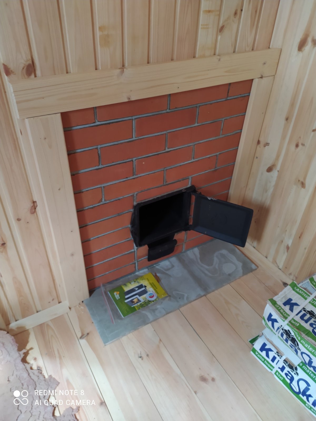 Wnętrze wykończone drewnem z murowanym kominem pieca, widoczna otwarta klapka paleniska, instrukcja obsługi i worki z kamieniami do pieca.