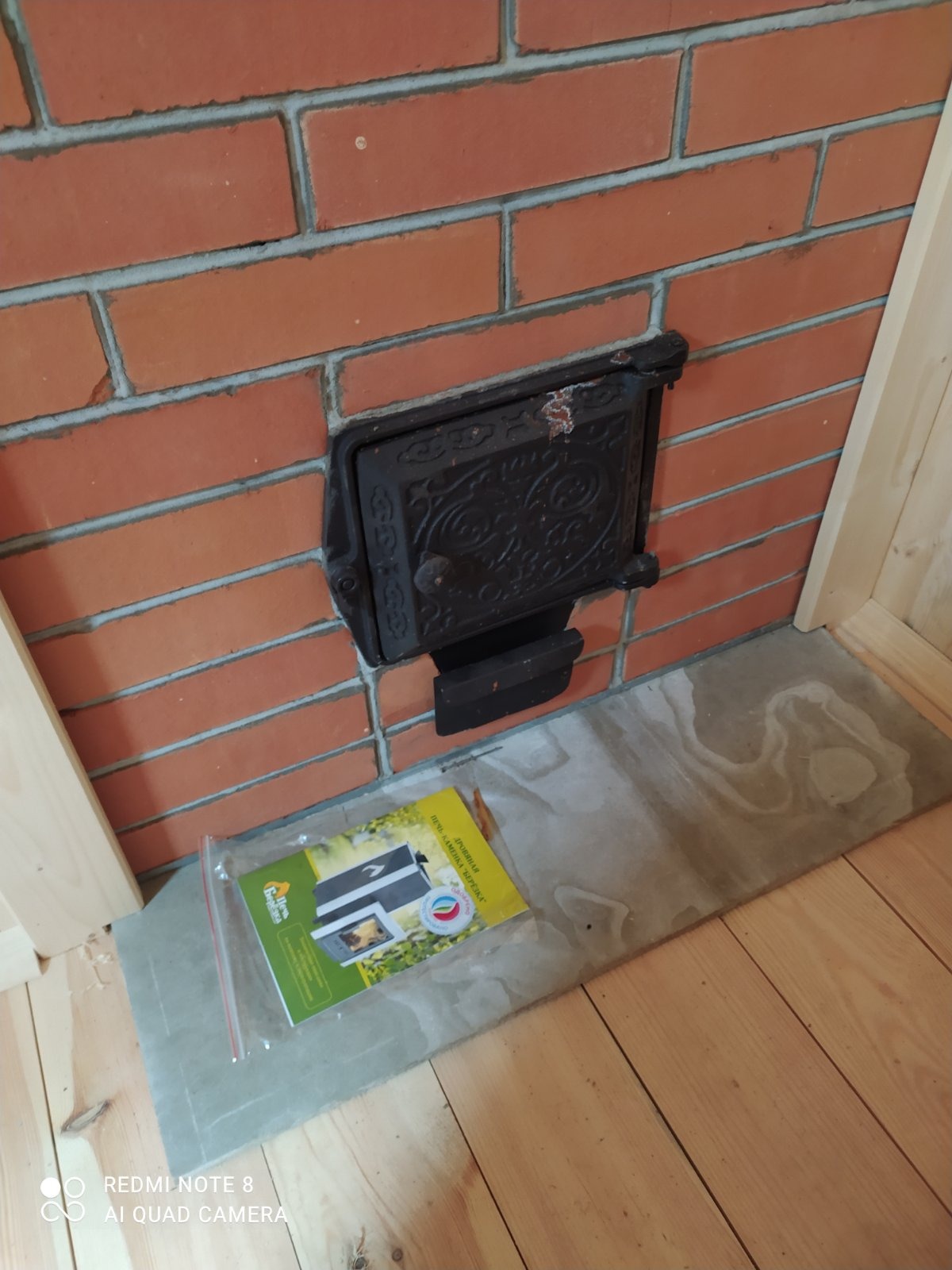 Fragment murowanej ściany z cegły z żeliwnymi drzwiczkami pieca, leżąca obok ulotka informacyjna w folii, widoczna drewniana podłoga i fragment ościeżnicy.